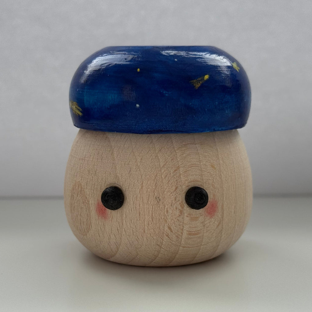 A small wooden toy figure with a round natural wood body, black painted eyes, pink cheeks, and a glossy dark blue cap decorated with tiny gold stars, set against a neutral gray background.