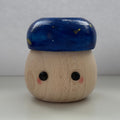A small wooden toy figure with a round natural wood body, black painted eyes, pink cheeks, and a glossy dark blue cap decorated with tiny gold stars, set against a neutral gray background.