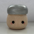 A small wooden character figurine with a round natural wood body, black dot eyes, pink cheek details, and a metallic silver cap, set against a neutral gray background.