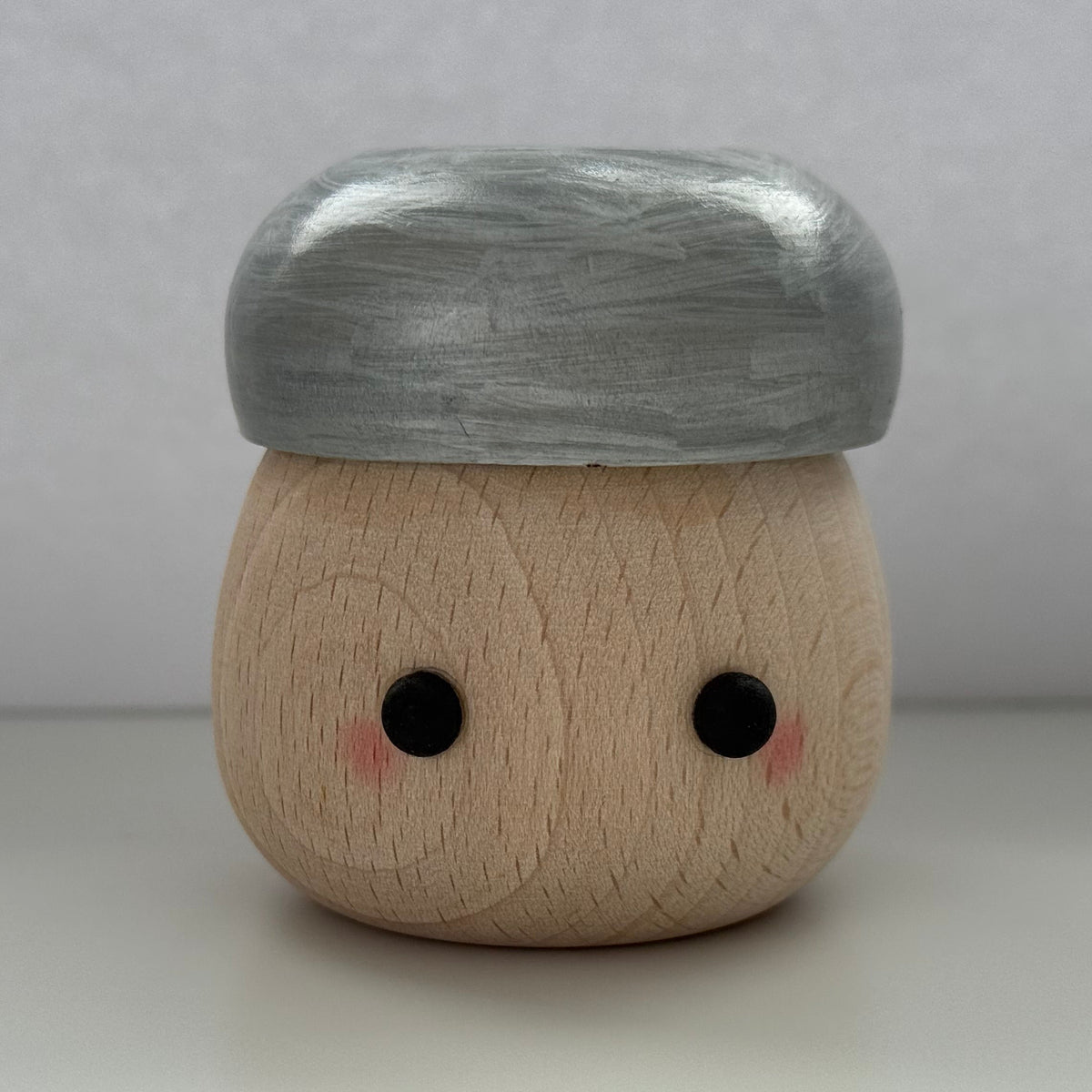 A small wooden character figurine with a round natural wood body, black dot eyes, pink cheek details, and a metallic silver cap, set against a neutral gray background.