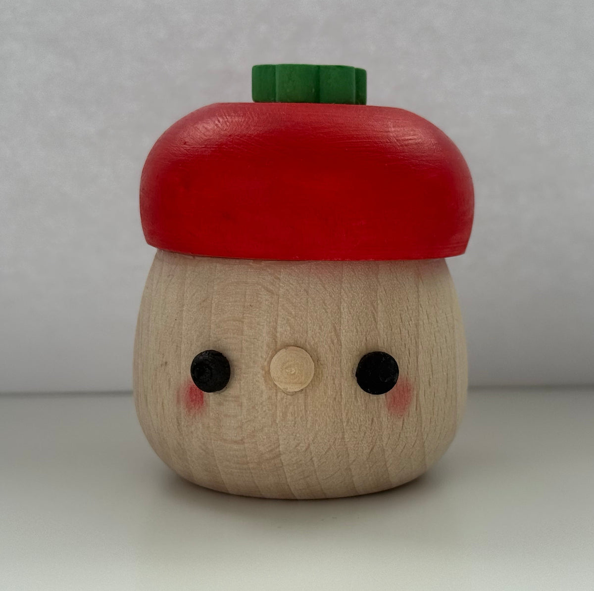 A small wooden acorn-shaped container with a red painted cap, a green top piece, and a cute face carved into the natural wood body, set against a neutral gray background.