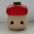 A small wooden acorn figurine with a red cap decorated with yellow seeds, a green stem, and a light natural wood body with black eyes and pink cheeks, set against a plain light gray background.