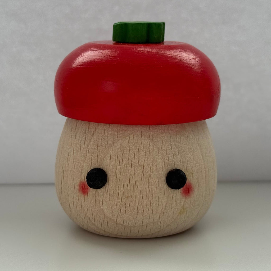 A small wooden acorn-shaped container or toy with a red painted cap, a natural wood body, black dot eyes, pink cheeks, and a green top piece, photographed against a plain light gray background.
