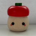 A small wooden acorn-shaped container or toy with a red painted cap, a natural wood body, black dot eyes, pink cheeks, and a green top piece, photographed against a plain light gray background.