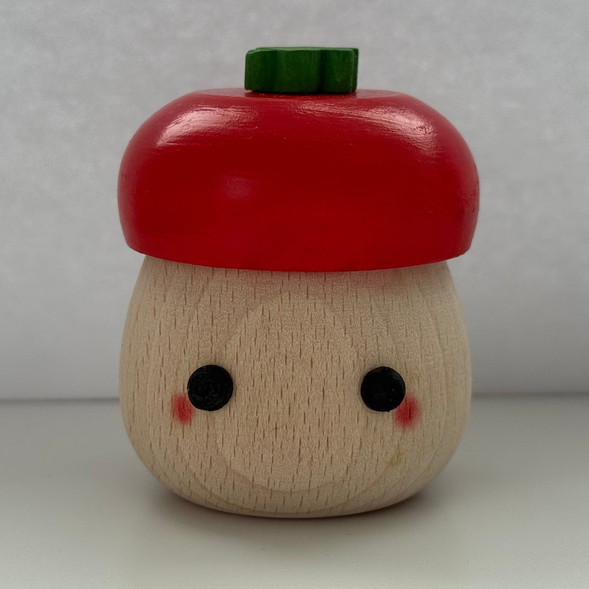 A small wooden acorn-shaped container or toy with a red painted cap, a natural wood body, black dot eyes, pink cheeks, and a green top piece, photographed against a plain light gray background.