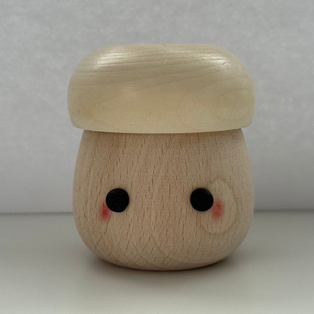 A small wooden mushroom-shaped figurine with a smiling face, featuring a light beige cap and a natural wood-toned body with black eyes and pink cheeks, set against a neutral gray background.