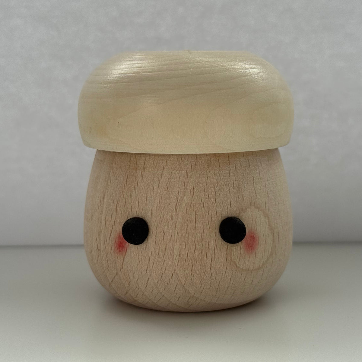 A small wooden mushroom-shaped figurine with a smiling face, featuring a light beige cap and a natural wood-toned body with black eyes and pink cheeks, set against a neutral gray background.