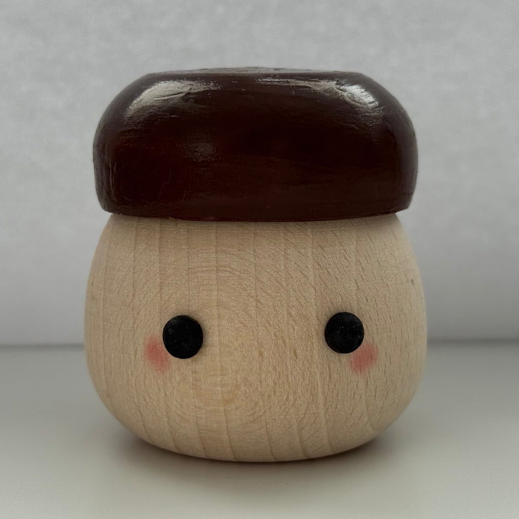 A small wooden acorn-shaped container or figurine with a smooth dark brown cap, natural light wood body, painted black eyes, and pink cheeks, centered against a plain light gray background.