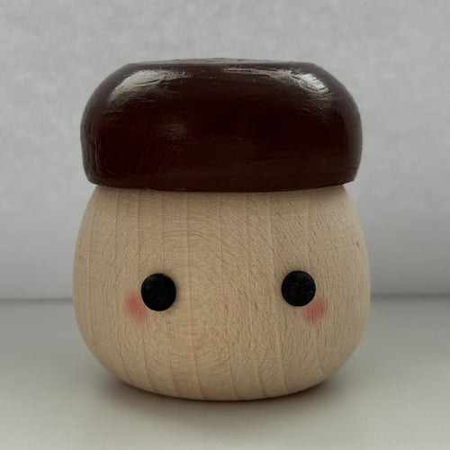 A small wooden acorn-shaped container or figurine with a smooth dark brown cap, natural light wood body, painted black eyes, and pink cheeks, centered against a plain light gray background.