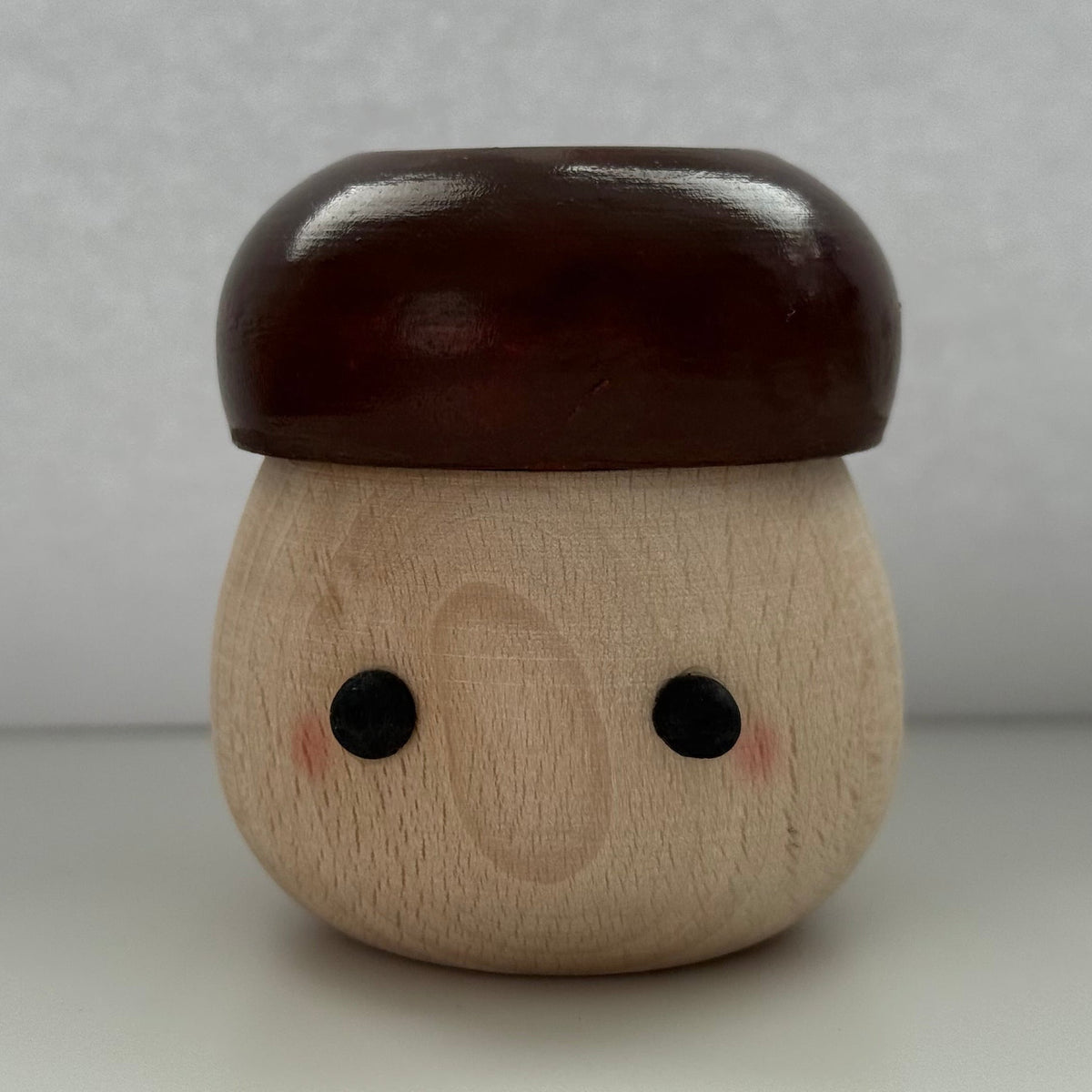 A small wooden acorn-shaped figurine with a glossy dark brown cap and a natural light wood body featuring simple black eyes and pink cheeks, set against a neutral gray background.