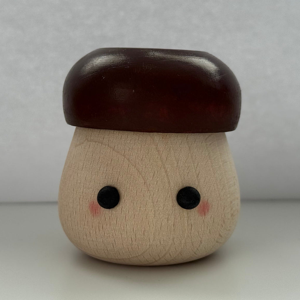 A small mushroom-shaped wooden toy or container with a light natural wood body, dark reddish-brown cap, and simple black eyes with pink cheeks, photographed against a plain light gray background.