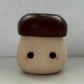 A small mushroom-shaped wooden toy or container with a light natural wood body, dark reddish-brown cap, and simple black eyes with pink cheeks, photographed against a plain light gray background.