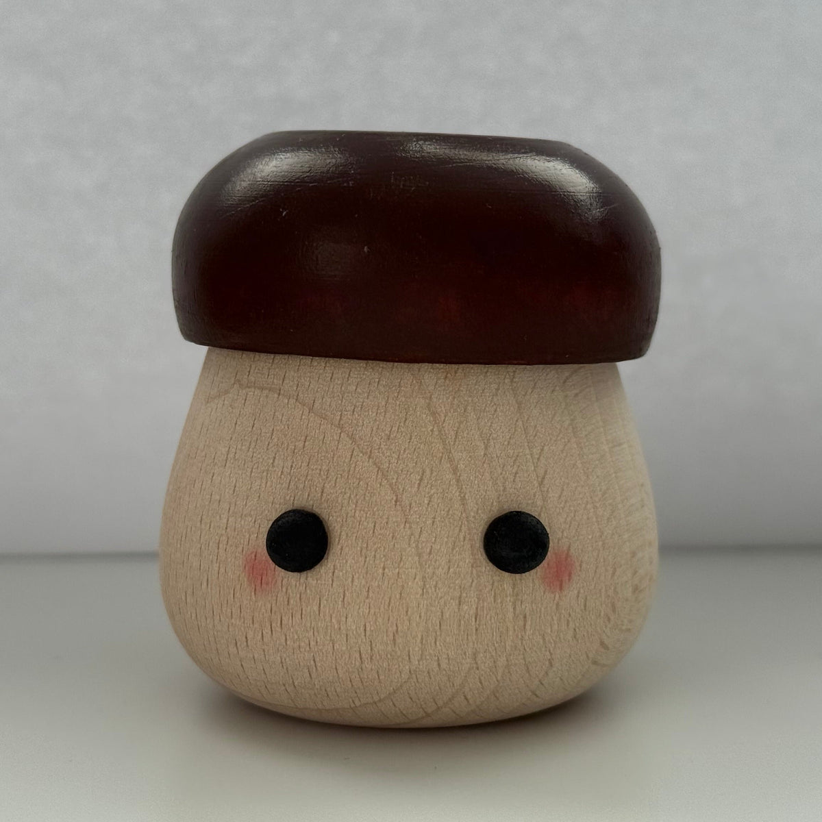 A small mushroom-shaped wooden toy or container with a light natural wood body, dark reddish-brown cap, and simple black eyes with pink cheeks, photographed against a plain light gray background.