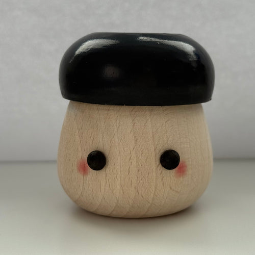 A small wooden toy figure with a rounded beige body, black painted eyes, pink cheeks, and a glossy black cap-like top, photographed against a plain light gray background.