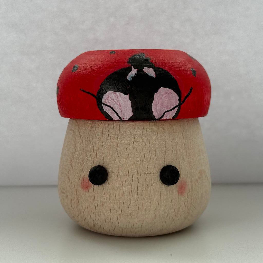 A small hand-painted wooden figurine shaped like a mushroom with a red cap, black and pink face details, and a natural wood base, set against a plain light gray background.