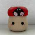 A small hand-painted wooden figurine shaped like a mushroom with a red cap, black and pink face details, and a natural wood base, set against a plain light gray background.