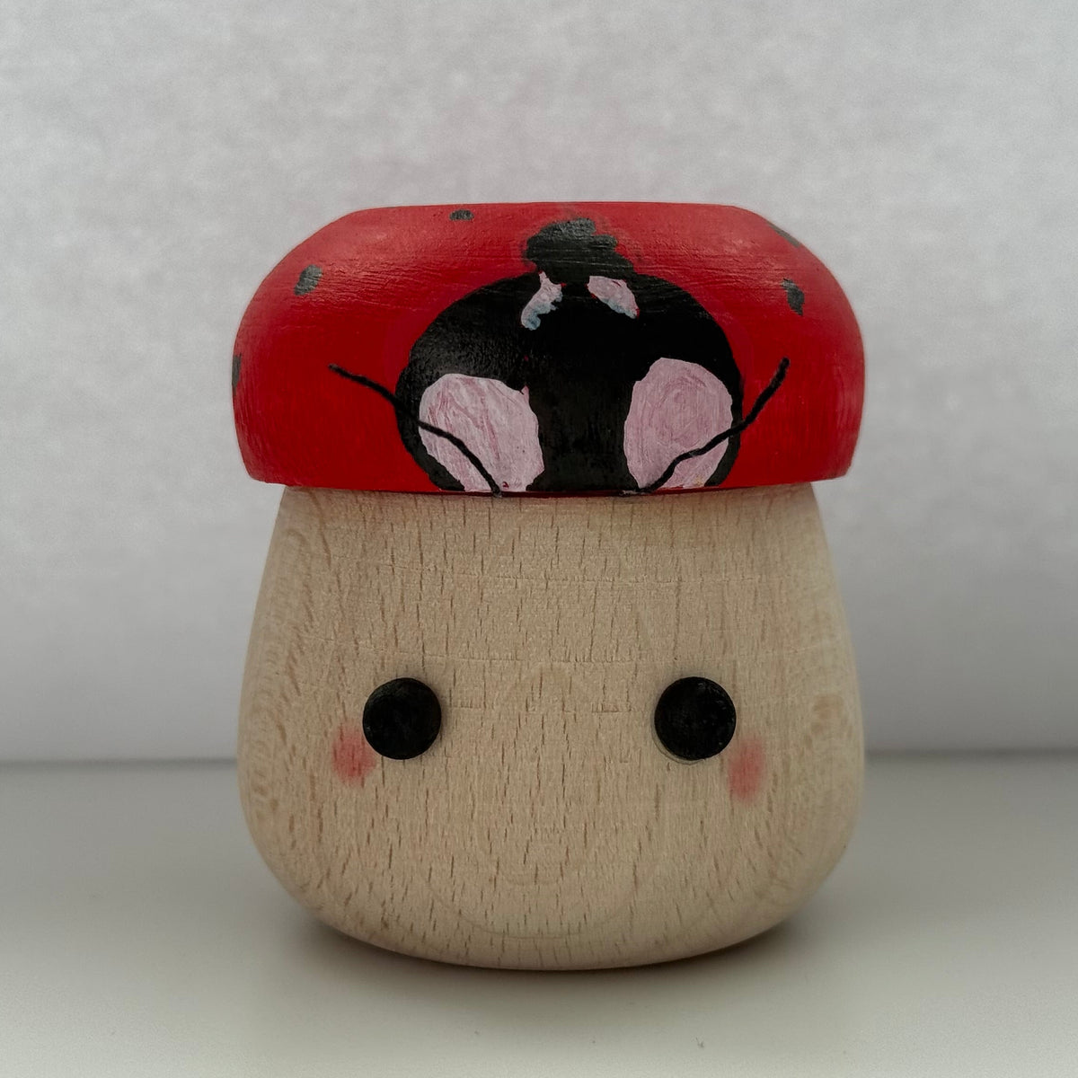 A small hand-painted wooden figurine shaped like a mushroom with a red cap, black and pink face details, and a natural wood base, set against a plain light gray background.
