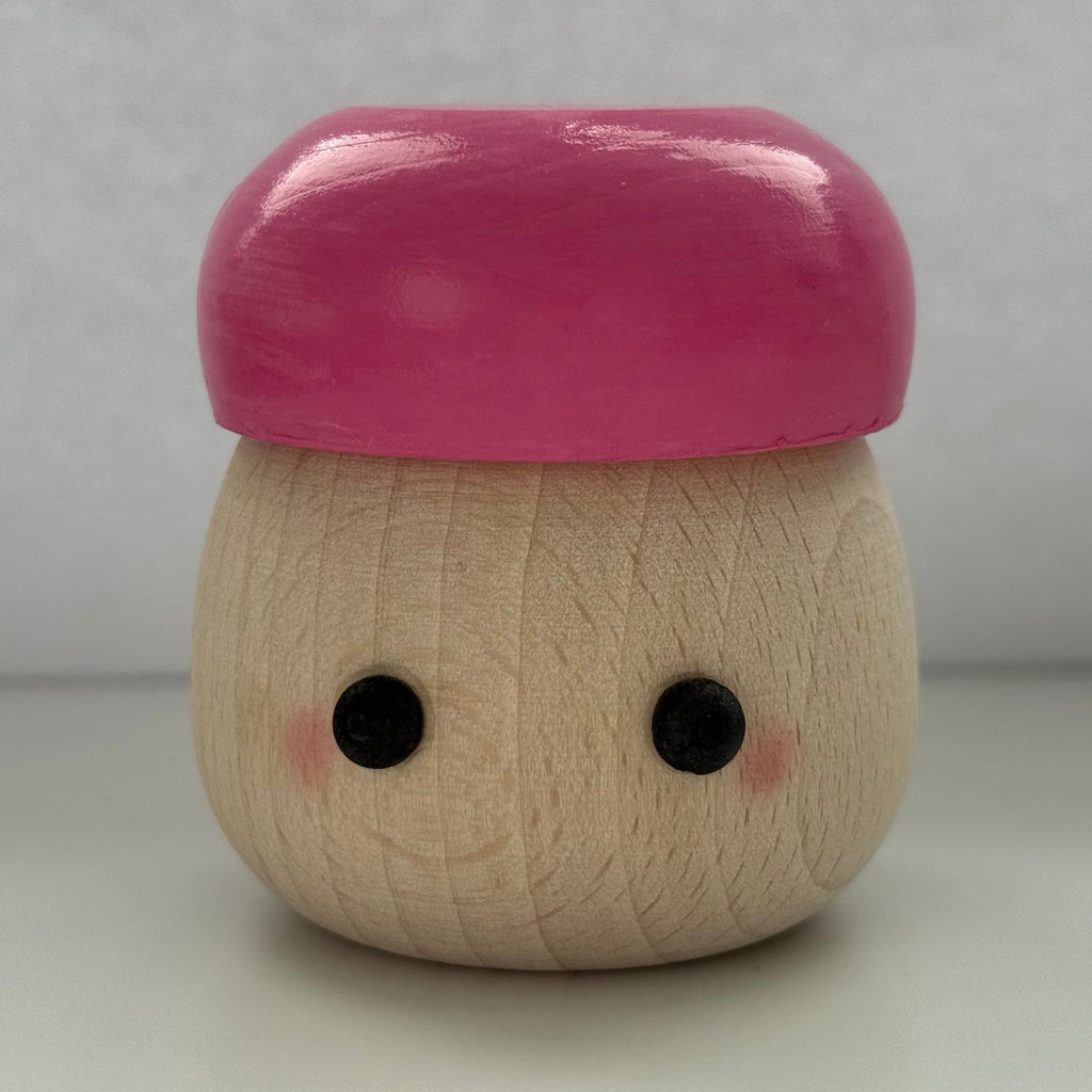 A small wooden toy figure with a smooth pink cap-like top and a natural wood body, featuring simple black eyes and pink cheeks, photographed against a plain light gray background.