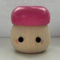 A small wooden toy figure with a smooth pink cap-like top and a natural wood body, featuring simple black eyes and pink cheeks, photographed against a plain light gray background.