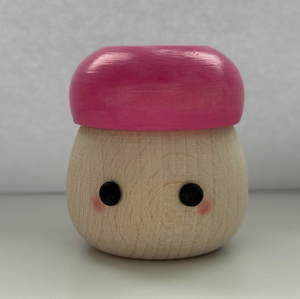 A small wooden toy figure with a round natural wood body, black dot eyes, pink cheeks, and a glossy pink cap, standing on a white surface against a light gray background.