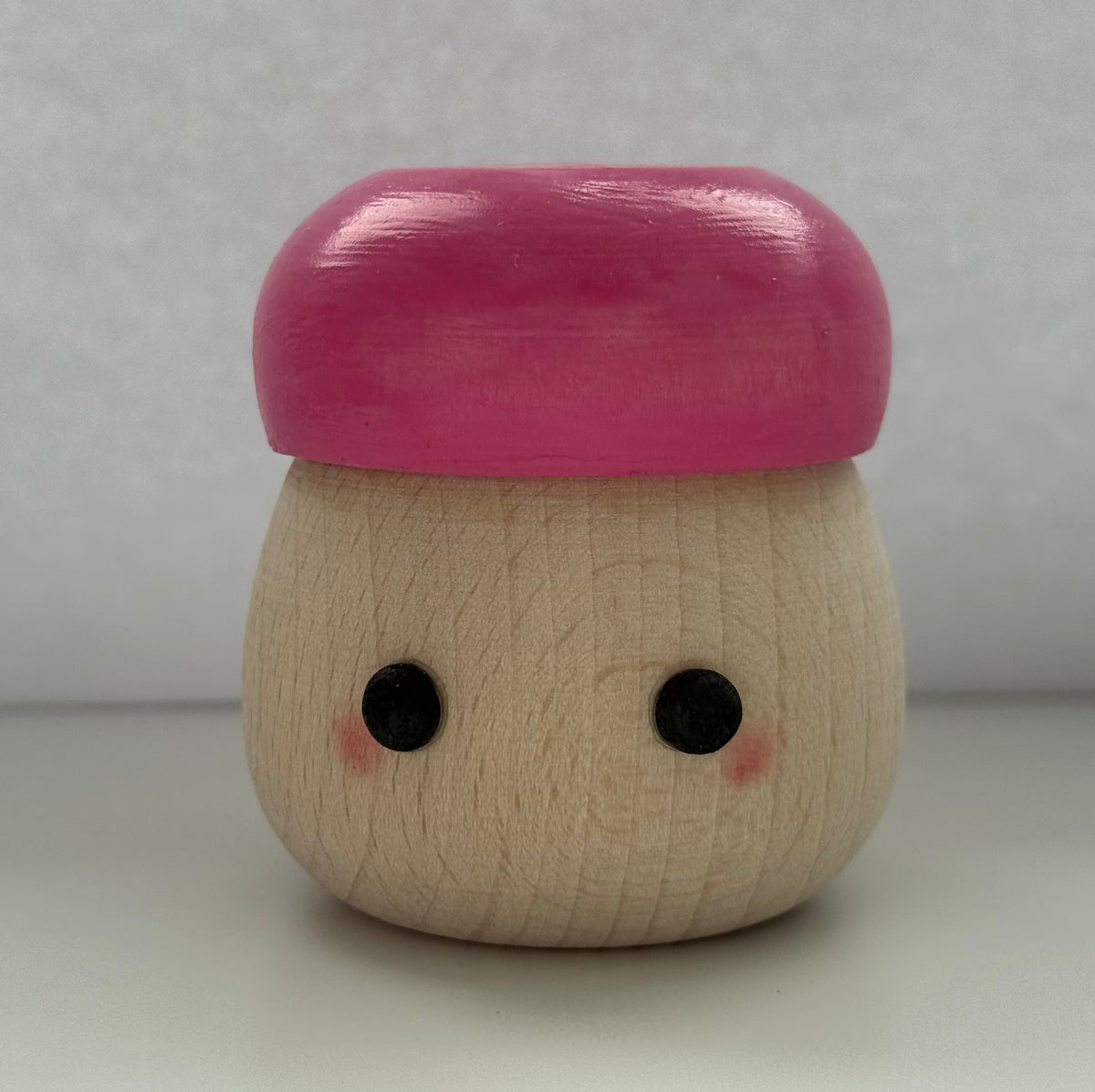 A small wooden toy figure with a round natural wood body, black dot eyes, pink cheeks, and a glossy pink cap, standing on a white surface against a light gray background.