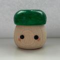 A small mushroom-shaped wooden toy or figurine with a glossy dark green cap and a natural light wood body featuring simple black eyes and pink cheeks, set against a plain light gray background.