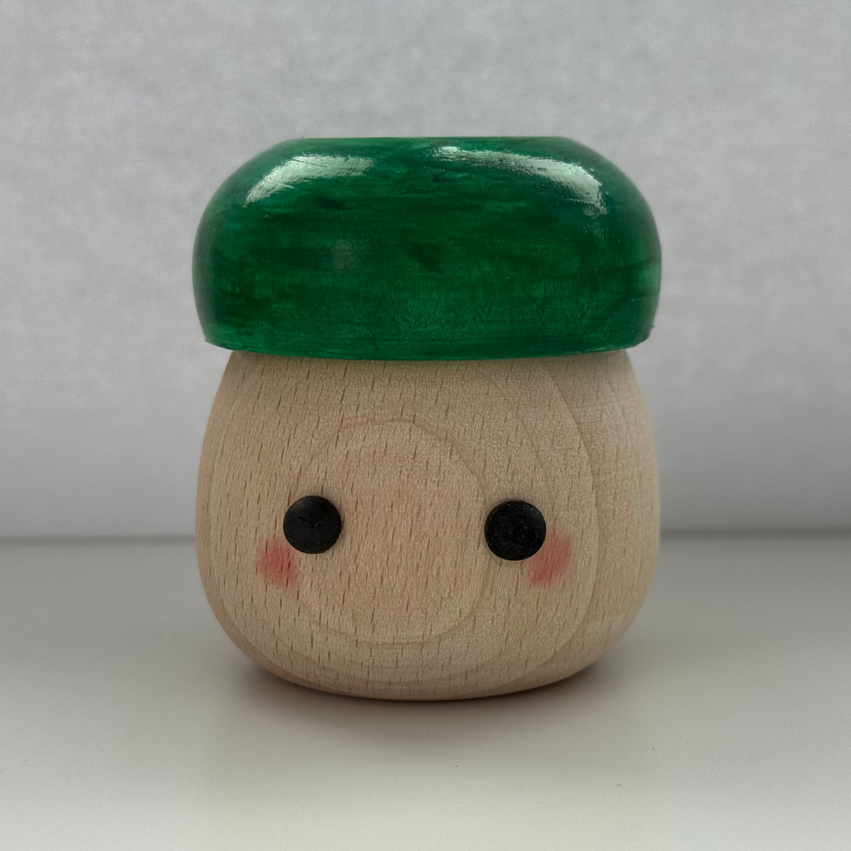 A small mushroom-shaped wooden toy or figurine with a glossy dark green cap and a natural light wood body featuring simple black eyes and pink cheeks, set against a plain light gray background.