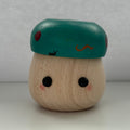A small wooden toy figure with a round natural wood body, black dot eyes, pink cheeks, and a teal cap decorated with red spots and a small orange mark, set against a plain light gray background.