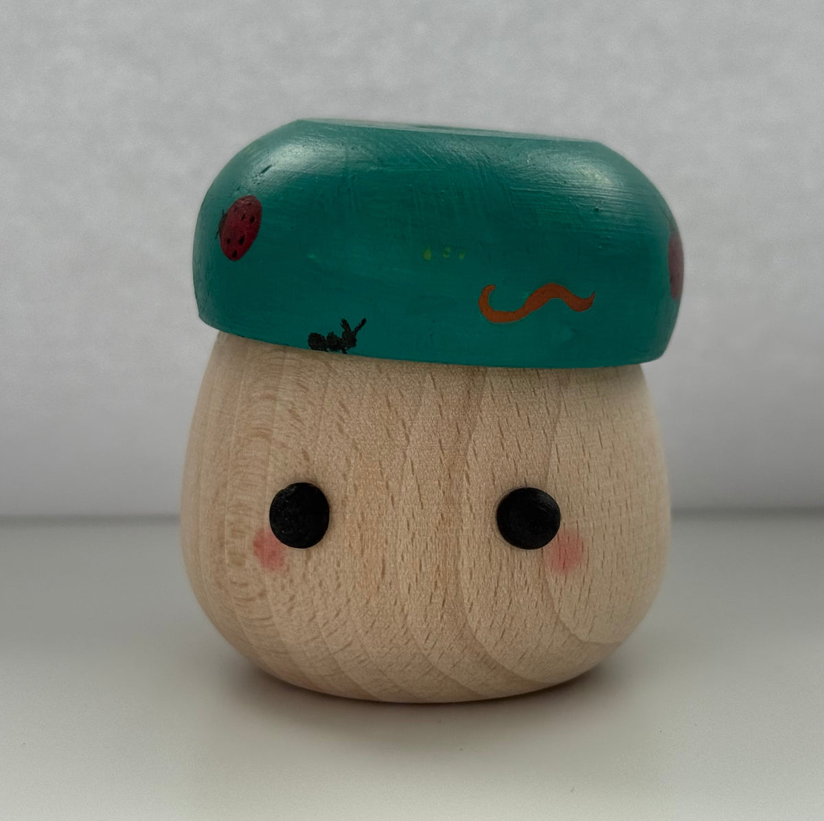 A small wooden toy figure with a round natural wood body, black dot eyes, pink cheeks, and a teal cap decorated with red spots and a small orange mark, set against a plain light gray background.